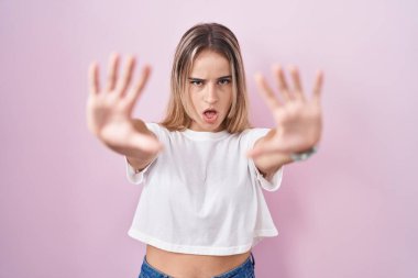 Young blonde woman standing over pink background doing stop gesture with hands palms, angry and frustration expression 