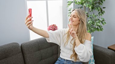 Gülümseyen genç bir kadın kapalı bir oturma odasında telefonuyla selfie çekiyor..