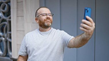 Kendine güveni tam beyaz bir adam, güneşli bir caddede akıllı telefonuyla eğlence dolu bir selfie çekerken gülümsüyor..