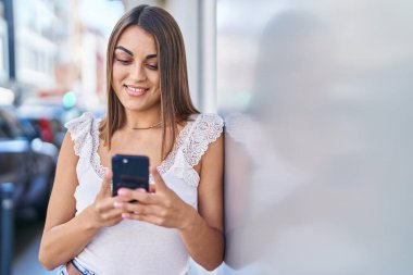 Genç, güzel, İspanyol bir kadın sokakta akıllı telefon kullanarak gülümsüyor.