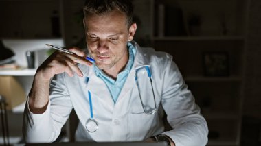 Steteskopla laboratuar önlüklü dalgın bir erkek doktor gece vakti hastane ofisindeki bilgisayardan hasta bilgilerini inceliyor..
