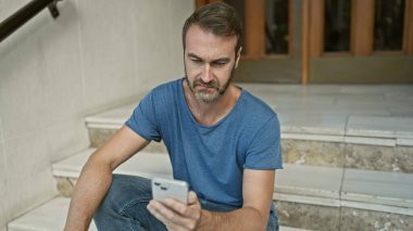 Sakallı yakışıklı İspanyol adam şehir basamaklarında otururken akıllı telefon kullanıyor..