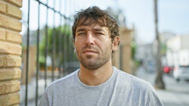 Ciddi bir ifadeyle kendine güvenen genç bir adam, bir sokak ortamında dikiliyor, konsantre ama rahat bir dış görünüşü canlandırıyor.