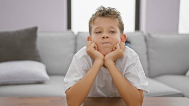 Sarışın çocuk masaya yaslanıyor ve evde düşünüyor.