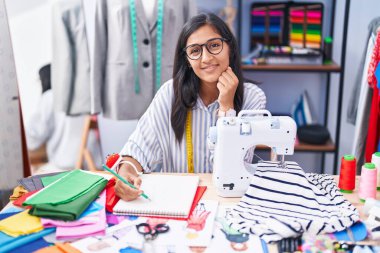 Young beautiful hispanic woman tailor smiling confident drawing on notebook at clothing factory