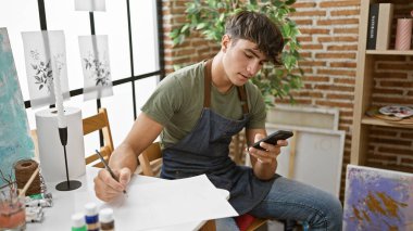 Sanat stüdyosunun içinde, tutkulu genç İspanyol asıllı genç, sanatsal ve öğrenme dalına dalmış, yaratıcı bir vızıltının ortasında akıllı telefona bakarken,