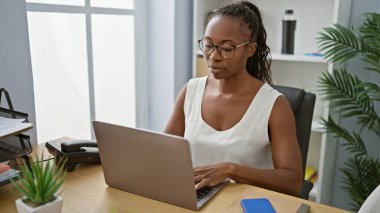 Parlak bir ofis ortamında dizüstü bilgisayar üzerinde çalışan odaklanmış Afrikalı kadın, profesyonellik ve konsantrasyonu somutlaştırıyor..