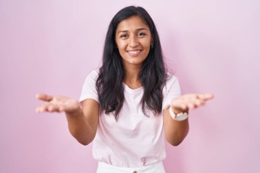 Young hispanic woman standing over pink background smiling cheerful offering hands giving assistance and acceptance. 