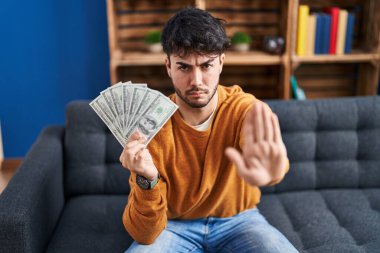 Sakallı İspanyol adam elinde 100 dolarlık banknotlarla elinde dur işareti ile ciddi ve kendinden emin bir ifadeyle savunma hareketi yapıyor. 