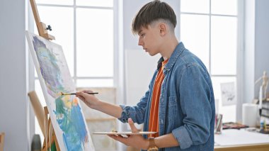 Odaklanmış genç bir adam parlak bir sanat stüdyosundaki tuvale resim yapar, yaratıcılığı ve konsantrasyonu somutlaştırır..