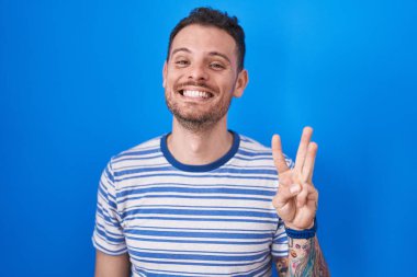 Young hispanic man standing over blue background showing and pointing up with fingers number three while smiling confident and happy. 