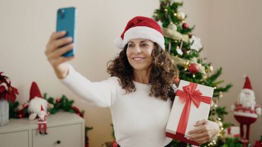 Orta yaşlı İspanyol kadın evde elinde Noel hediyesi olan akıllı telefonuyla selfie çekiyor.