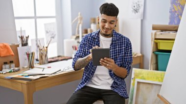 Yakışıklı genç Latin ressam dokunmatik ped kullanıyor, hareketli sanat stüdyosunda tuvale resim yaparken gülümsüyor, yaratıcılığı ve tutkuyu somutlaştırıyor..