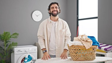 Genç İspanyol adam gülümsüyor. Çamaşır odasındaki masayı ütülüyor.