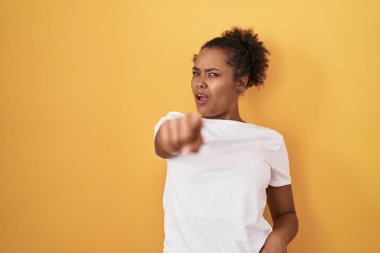 Young hispanic woman with curly hair standing over yellow background pointing displeased and frustrated to the camera, angry and furious with you 