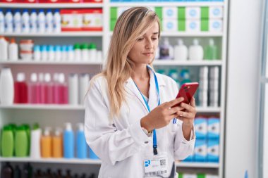 Young blonde woman pharmacist using smartphone working at pharmacy