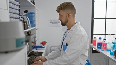 Laboratuvar önlüğü giyen beyaz sakallı genç adam parlak, modern bir laboratuvarda bilgisayar üzerinde çalışıyor.
