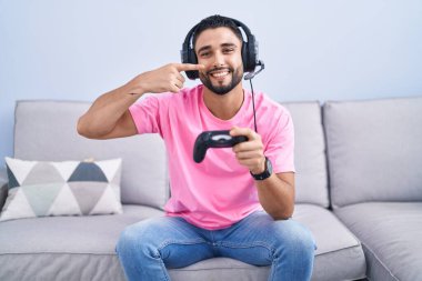 İspanyol asıllı genç adam video oyunu oynuyor kanepeye oturmuş, parmaklarını yüzüne ve burnuna göstererek neşeli bir şekilde gülümsüyor. güzellik konsepti 
