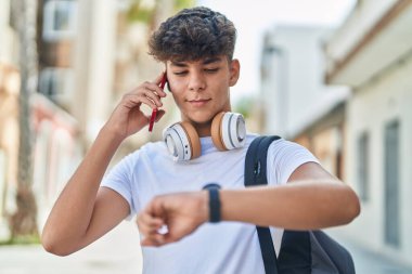 Young hispanic teenager student talking on smartphone looking watch at street
