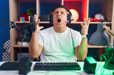 Young hispanic man playing video games crazy and mad shouting and yelling with aggressive expression and arms raised. frustration concept. 