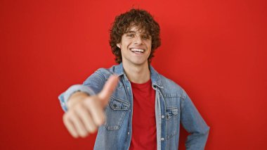 Kıvırcık saçlı neşeli bir İspanyol adam kırmızı arka planda izole edilmiş, sıradan kırmızı bir tişörtün üzerine kot ceket giyiyor..