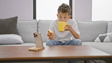 Sevimli sarışın çocuk, akıllı telefondan video seyretmeye dalmış, kahvaltılık kek atıştırmalıklarını evde, kanepede oturarak yiyor.
