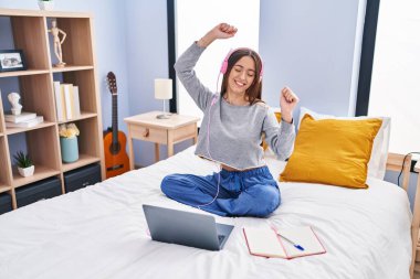 Young beautiful hispanic woman student listening to music studying on bed at bedroom