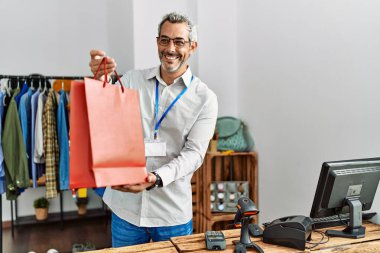 Orta yaşlı, gri saçlı, mağazada alışveriş çantası tutan bir dükkan asistanı.