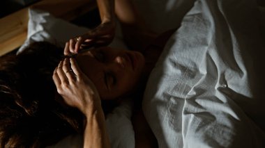 Yatakta baş ağrısı olan genç beyaz bir kadın, bir ev yatak odasında yatarken rahatsız edici bir his yaratıyor..
