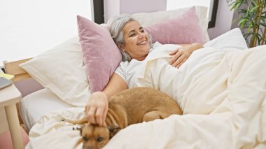 Gülümseyen orta yaşlı bir kadın evcil köpeğiyle yatakta rahatlatıcı bir andan zevk alır, evde rahatlık ve arkadaşlık hissi yakalar..