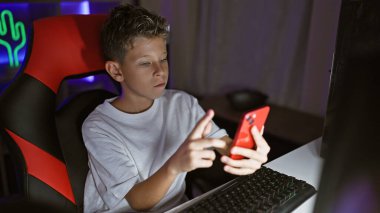 Sevimli sarışın çocuk flaması, karanlık, neon ışıklı bir odada oyun oynamaya odaklanmış, bir bilgisayar ve akıllı telefon arasında dinamik bir şekilde hokkabazlık yapıyor, dijital yayın dünyasına dalmış..