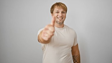 Sakallı ve dövmeli neşeli genç bir adam beyaz bir arka plana karşı baş parmak işareti yapıyor..