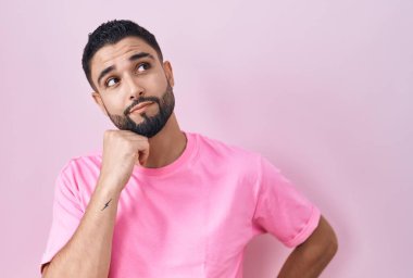 Elleri çenesinde pembe arka planda duran İspanyol genç adam soru sormayı, düşünceli ifadeyi düşünüyor. Düşünceli bir yüzle gülümsüyor. şüphe kavramı. 