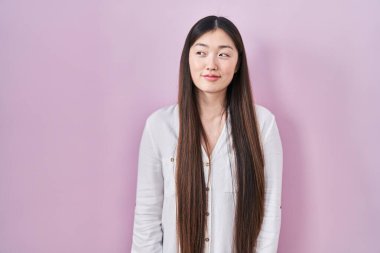 Pembe arka planda duran Çinli genç kadın gülümsüyor, yan tarafa bakıyor ve düşünüyor.. 