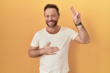 Sarı arka planda sakallı orta yaşlı bir adam gülümsüyor, elleri göğsünde ve parmakları havada küfür ediyor, sadakat yemini ediyor. 