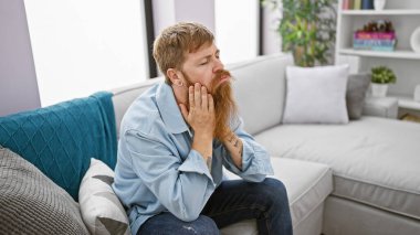 Depresyondaki genç kızıl saçlı adam, stresli ve yalnız, evdeki kanepede oturuyor, zihinsel kriz ve endişeyle boğuşuyor.