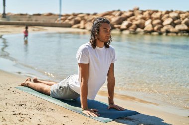 Young hispanic man doing yoga exercise at beach