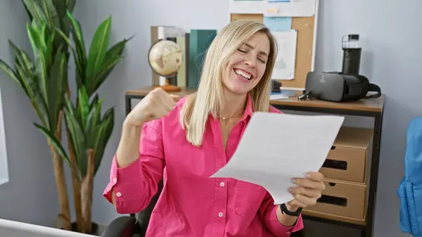 現代のオフィスの設定で紙を読みながらピンクのシャツを着ている陽気なブロンドの女性 — ストック写真