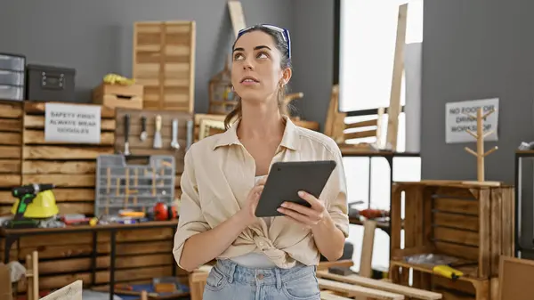 Attractive young hispanic woman carpenter looking around her carpentry workplace, skillfully using a touchpad amidst the timber and woodwork, bringing together beauty and industry