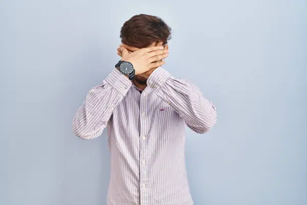 Arab man with beard standing over blue background covering eyes and mouth with hands, surprised and shocked. hiding emotion