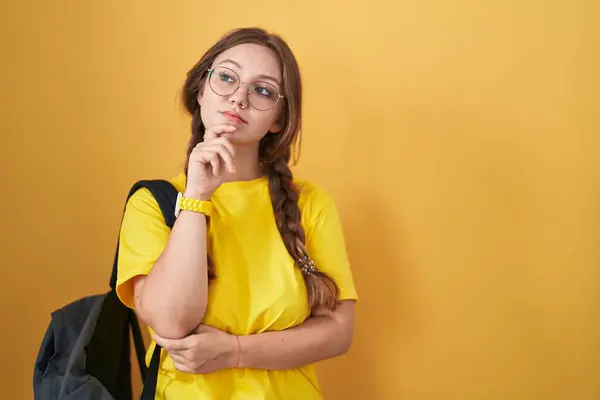 Young caucasian woman wearing student backpack over yellow background with hand on chin thinking about question, pensive expression. smiling with thoughtful face. doubt concept.