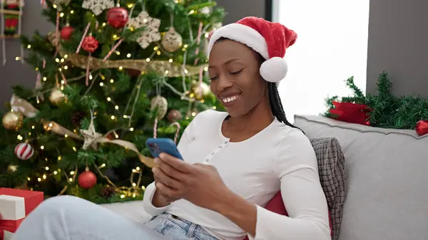 African american woman using smartphone sitting by christmas tree at home