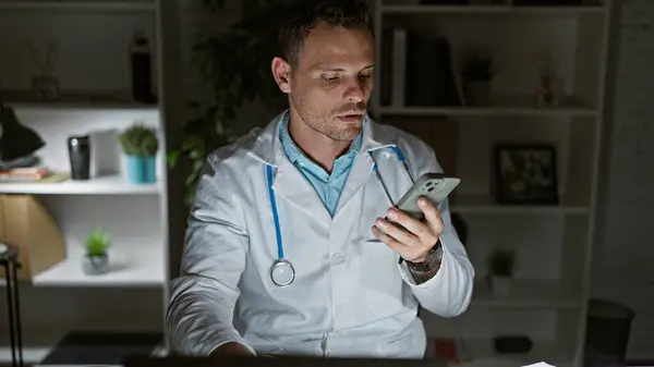 Hispanic man in lab coat scrutinizing smartphone indoors, portraying a clinic professional during a night shift.