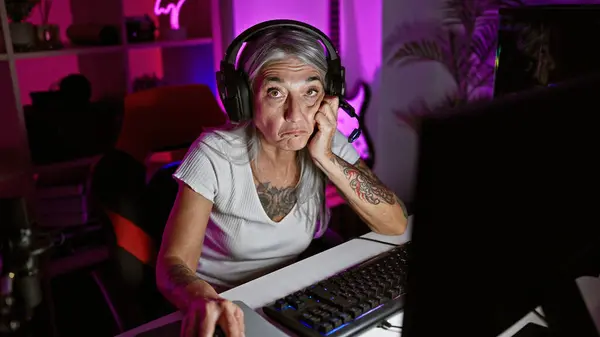 Mature, grey-haired woman gamer, in serious gaming mode, streaming live in the heart of the night from her cozy home gaming room, sporting headphones and a mic
