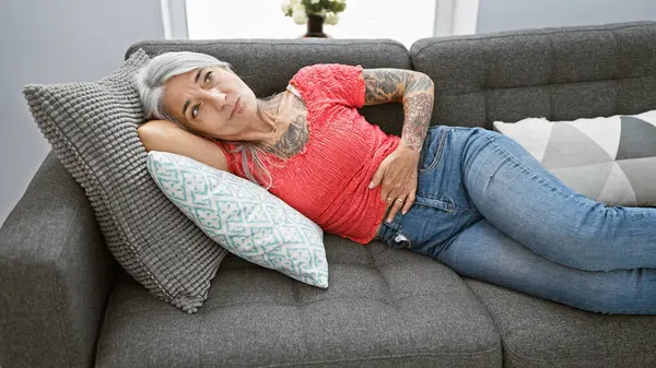 Worried middle-aged woman, gracefully grey-haired, suffering severe stomachache while resting, unhappily lying on her living room sofa indoors.