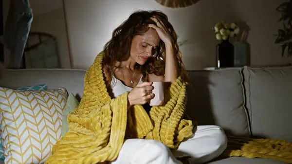A pensive woman in a cozy living room holds a mug while wrapped in a yellow blanket, conveying solitude and contemplation.