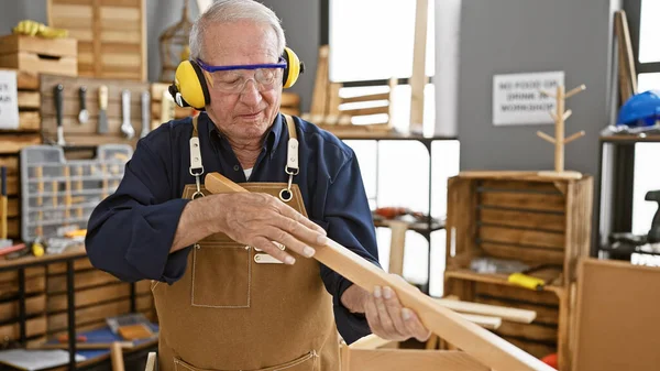 有弹性的老人是一位经验丰富的木匠 他坚定地站在自己的木工车间里 戴着安全玻璃 手里拿着一块刚锯好的木板 — 图库照片