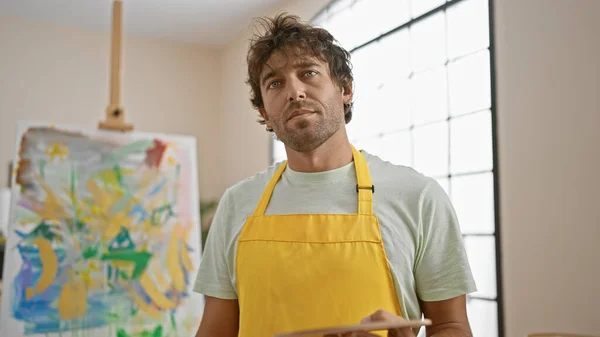 Handsome bearded man artist in studio with yellow apron creates abstract painting indoors