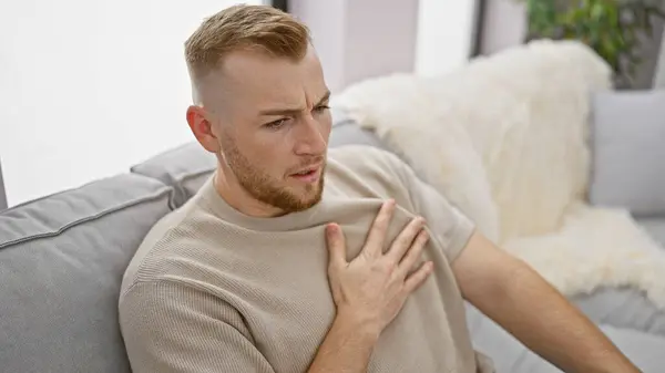 stock image Caucasian bearded man experiencing chest pain while sitting on a couch indoors.