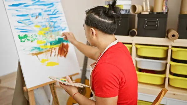 Serious young chinese man artist, relaxed yet concentrated, draws at his studio. handsome with pigtail hairstyle and apron, mastering the art on canvas with paintbrushes, embracing creativity.